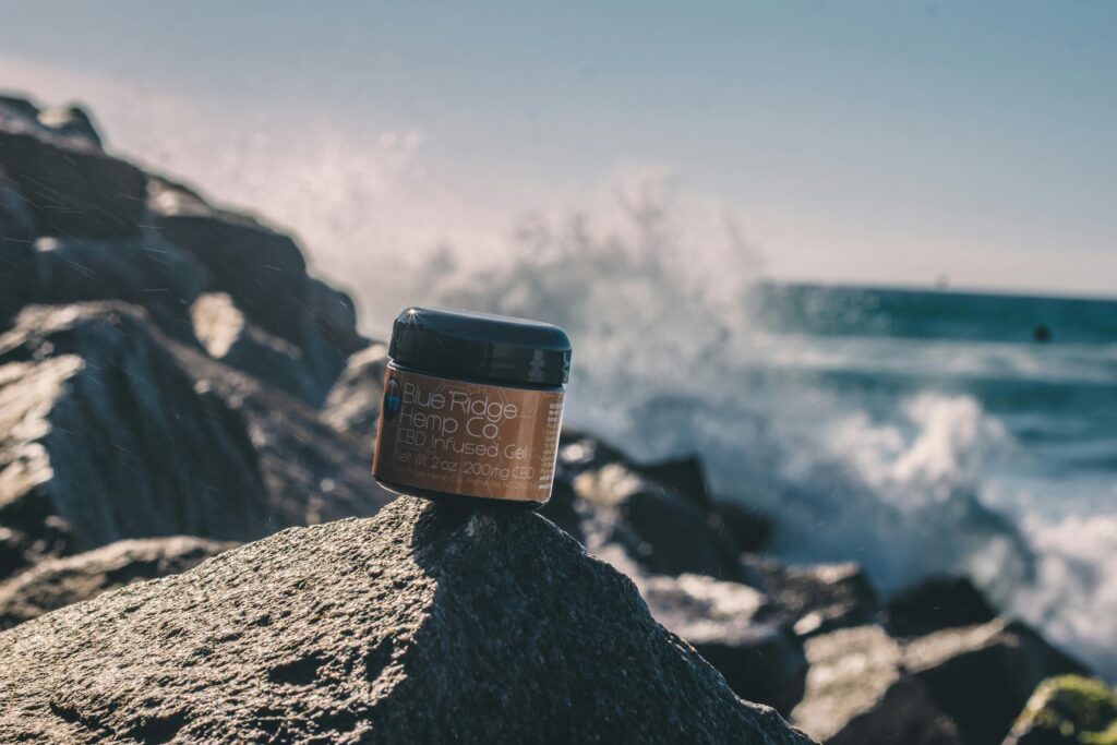 Ambalaj ve Ürün Tasarımı Close-up of CBD infused gel jar on rocks by the sea, showcasing product against a scenic ocean backdrop with splashing waves.